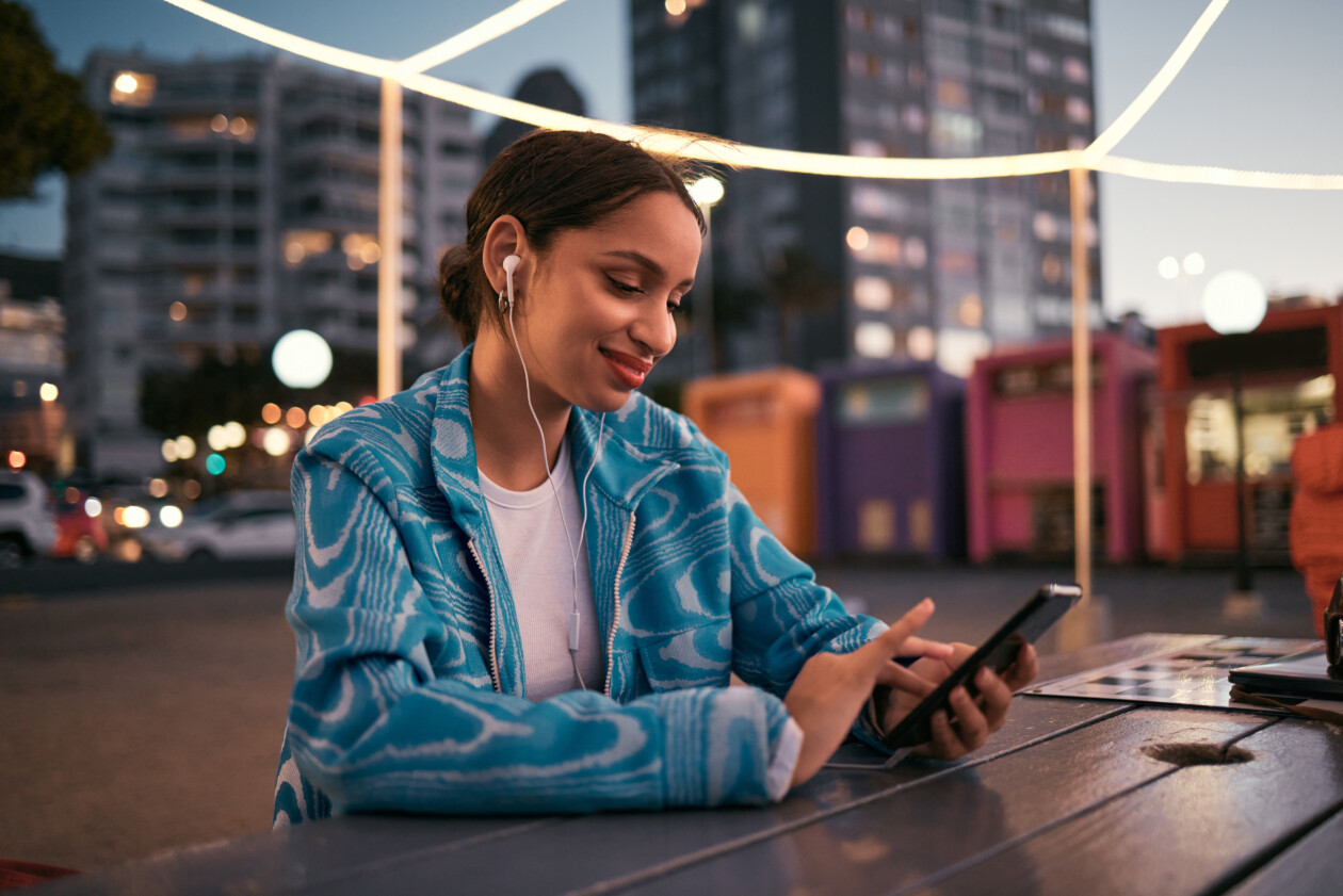 Happy and trendy woman browsing on phone while wearing earphones and listening to music while sitting at outdoor cafe in night city. happy woman streaming subscription app or making video call